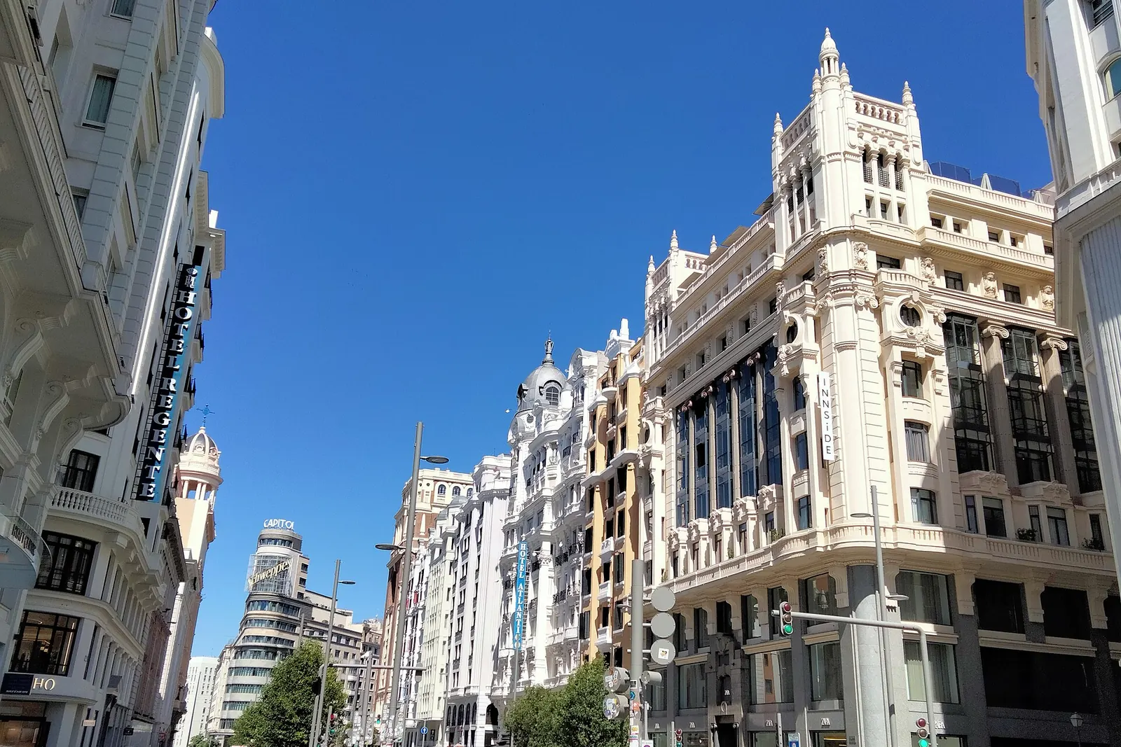 Edifici bianchi e color crema costeggiano un tratto soleggiato della Gran Vía di Madrid sotto un cielo blu intenso, con il Palazzo Capitol sullo sfondo